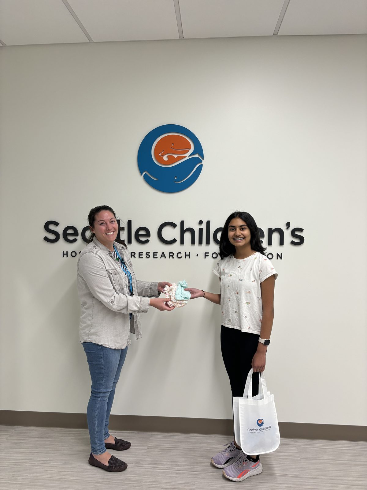 Monica handing a bundle of knitted baby hats to a Seattle Children’s nurse, in front of the hospital’s logo