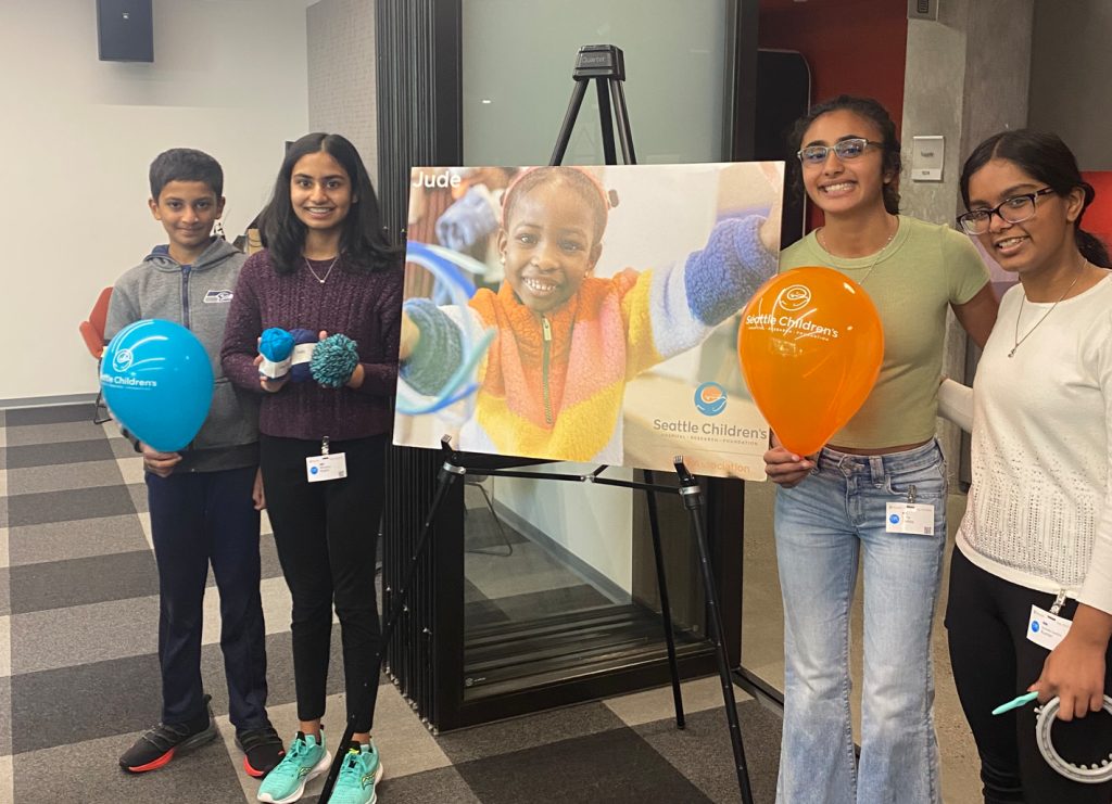 Guild members standing beside a Seattle Children’s display with balloons and knitted items at a campus event