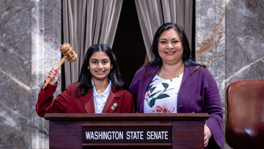 Monica standing at the rostrum in the Washington State Senate chamber, holding a gavel, beside Senator Manka Dhingra